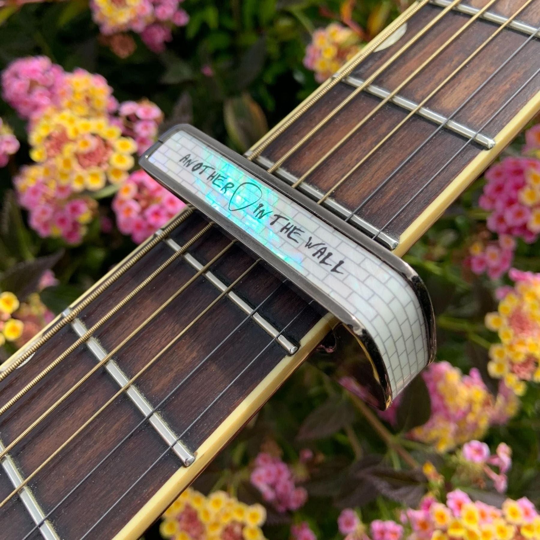 Thalia black-chrome capo with The Wall of Pearl inlay — angled fretboard view with flowers in the background — Slide 6 of 8